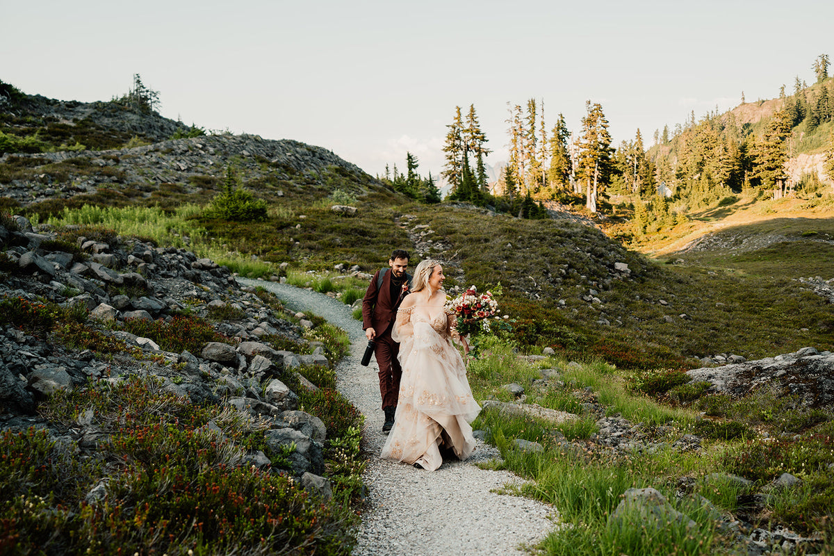 A bride and a groom laugh as they walk through a stony path through a mountain trail.
