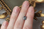 A natural cushion cut steely gray spinel available to add as a center stone for your heirloom setting at The North Way Studio. A steely blueish gray reminiscent of a stormy day. The spinel is shown resting on a woman's fingers for size reference.