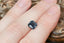 A natural emerald cut teal blue opalescent sapphire available to add as a center stone for your heirloom ring setting at The North Way Studio. A faintly opalescent blue with hints of teal within its depths, this stone is shown resting on a woman's fingers for size reference.