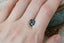 A natural emerald cut teal blue opalescent sapphire available to add as a center stone for your heirloom ring setting at The North Way Studio. A faintly opalescent blue with hints of teal within its depths, this stone is shown resting on a woman's hand for size reference.