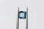 A natural emerald cut teal blue opalescent sapphire available to add as a center stone for your heirloom ring setting at The North Way Studio. A faintly opalescent blue with hints of teal within its depths, this stone is shown on a white background, tilted to the side and held by tweezers.