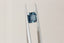 A natural emerald cut teal blue opalescent sapphire available to add as a center stone for your heirloom ring setting at The North Way Studio. A faintly opalescent blue with hints of teal within its depths, this stone is shown titled to the side and held by tweezers.