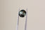 A natural round green teal sapphire available to add as a center stone for your heirloom setting at The North Way Studio. Shifting hues of teals, blues, and greens. The sapphire is shown held up by tweezers.