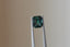 A natural emerald cut teal sapphire available to add as a center stone for your heirloom setting at The North Way Studio. A deeper forest teal hue. The sapphire is shown held up with tweezers.
