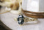 A customizable fantasy solitaire engagement ring setting with small brilliant cut diamonds set into leaf prongs. Shown in reclaimed 14 karat yellow gold with an oval dark gray spinel. Elven fairytale engagement ring designed by The North Way Studio.
