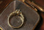 This fantasy customizable engagement ring setting crafted by The North Way studio features vines of reclaimed gold unfurling about genuine diamond dewdrops and a center stone chosen by the customer. This custom engagement ring is shown in 14 karat gold with a gray spinel center resting atop an antique mirror and viewed from above. Elven engagement ring.