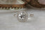 This antique inspired heirloom setting crafted by The North Way Studio features leaves carved in reclaimed gold, a halo of natural brilliant cut round diamonds, and two round natural diamond side stones strewn about a unique center stone. This custom medieval fantasy engagement ring is shown in 14 karat white gold with a round diamond center sitting facing the viewer on a marble table.