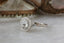 This fantasy antique inspired heirloom setting crafted by The North Way Studio features leaves carved in reclaimed gold, a halo of natural brilliant cut round diamonds, and two round natural diamond side stones strewn about a unique center stone. This customizable engagement ring is shown in 14 karat white gold with a round diamond center facing the viewer's left on a marble table.
