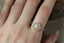 This fairytale antique inspired heirloom engagement ring setting crafted by The North Way Studio features leaves carved in reclaimed gold, a halo of natural brilliant cut round diamonds, and two round natural diamond side stones strewn about a unique center stone. This custom fantasy alternative engagement ring is shown in 14 karat yellow gold with a round diamond center and worn on the index finger of a hand with long nails.