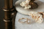 This fantasy heirloom setting crafted by The North Way Studio features leaves carved in reclaimed gold, a halo of natural brilliant cut round diamonds, and two round natural diamond side stones strewn about a unique center stone. This custom elven antique inspired engagement ring is shown in 14 karat gold with a round diamond center sitting facing the viewer's right on a marble table.