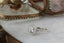 This antique inspired fantasy heirloom setting crafted by The North Way Studio features leaves carved in reclaimed gold, a halo of natural brilliant cut round diamonds, and two round natural diamond side stones strewn about a unique center stone. This custom fairytale engagement ring is shown in 14 karat white gold with a round diamond center sitting facing the viewer's left on a marble table in front of a glass antique jewelry box.