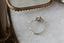 This antique inspired heirloom setting crafted by The North Way Studio features leaves carved in reclaimed gold, a halo of natural brilliant cut round diamonds, and two round natural diamond side stones strewn about a unique center stone. This custom medieval fantasy engagement ring is shown in 14 karat white gold with a round diamond center sitting on a marble table in front of a glass antique jewelry box and viewed from above.