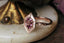 This custom fantasy engagement ring setting crafted by The North Way Studio features carved leaves placed amongst a golden shield and a unique center stone of choice. This medieval inspired alternative solitaire engagement ring is shown in reclaimed 14 karat rose gold with a pink garnet center.