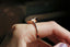 This custom solitaire engagement ring setting crafted by The North Way Studio features unfurling, carved leaves placed amongst a golden shield and a unique center stone of choice. This fantasy elf inspired alternative engagement ring is shown here in reclaimed 14 karat rose gold with a pink garnet center and worn on the index finger of a hand showing the side profile of the setting.