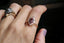 This custom elven engagement ring setting crafted by The North Way Studio features unfurling, carved leaves placed amongst a golden shield and a unique center stone of choice. This fantasy solitaire engagement ring is shown here in reclaimed 14 karat rose gold with a pink garnet center and worn on the index finger of a hand.