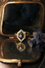 This custom fantasy elven style ring setting crafted by The North Way Studio features carved leaves placed amongst a golden shield and a unique center stone of choice. This solitaire engagement ring is shown here in reclaimed 14 karat yellow gold with a natural purple spinel hexagon center resting on an antique glass jewelry box beside a dried flower.