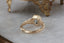 A blue green sapphire sits in the middle of this engagement ring setting designed by The North Way Studio shown resting on a marble table facing away from the viewer. A halo of diamonds surrounds the center stone and whittled bark textures and leaves are carved into the 14 karat gold.
