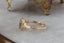 A blue green sapphire sits in the middle of this engagement ring setting designed by The North Way Studio shown resting on a marble table facing the viewer's left. A halo of diamonds surrounds the center stone and whittled bark textures and leaves are carved into the 14 karat gold.