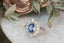 An antique inspired flower engagement ring with a large blue sapphire and natural diamond halo in 18 karat white gold with hand-carved leaves and bark texture. A ready to ship ring crafted by The North Way Studio in collaboration with Claire Pettibone.