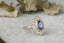 An antique inspired flower engagement ring with a large blue sapphire and natural diamond halo in 18 karat white gold with hand-carved leaves and bark texture. A ready to ship ring crafted by The North Way Studio in collaboration with Claire Pettibone.