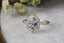 This heirloom engagement ring setting crafted by The North Way Studio features a shield halo of diamonds, engraved walls, and hand-carved leaves all strewn about a unique center stone of choice. This fantasy antique inspired engagement ring is shown in 14 karat white gold with a natural pale gray spinel center stone.