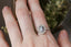 This heirloom engagement ring setting crafted by The North Way Studio features a shield halo of diamonds, engraved walls, and hand-carved leaves all strewn about a unique center stone of choice. This fantasy antique inspired engagement ring is shown in 14 karat white gold with a natural pale gray spinel center stone and worn on the index finger of a hand.