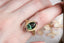 A one-of-a-kind enchanted garden engagement ring featuring a large forest green oval sapphire center with petite accent sapphires of mismatched blush and berry tones. Set in a lush 18 karat yellow gold with hand-carved leaves and a rustic finish. This luxury fantasy ring is designed by The North Way Studio in collaboration with Claire Pettibone and is ready to ship. Shown on a woman's hand.
