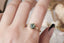 Vintage inspired enchanted garden engagement ring in teal green round sapphire, natural diamond, and 18 karat yellow gold. This ready to ship three stone engagement ring is shown on a woman's hand.