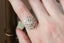 An antique inspired flower diamond halo engagement ring. This fairy garden engagement ring features a large natural gray diamond center with a diamond halo and hand-carved leaf and bark texture in 18 karat white gold. Ready to ship engagement ring crafted by The North Way Studio in collaboration with Claire Pettibone. Shown on a hand.