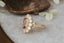 A flower engagement ring featuring a large natural pink spinel, diamond halo, marquise sapphire leaves, and hand carved bark texture in 18 karat yellow gold. An antique inspired fairy garden engagement ring. Ready to ship, crafted by The North Way Studio in collaboration with Claire Pettibone.