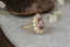 A flower engagement ring in natural pink spinel with diamond halo and sapphire leaves, hand carved leaves and bark texture in 18 karat yellow gold.