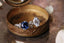This custom heirloom engagement ring setting designed and crafted by The North Way Studio features delicate leaves carved in reclaimed gold and a cluster of diamond dew drops spun about your choice of center gemstone. Two variations of this fantasy engagement ring are shown resting in an antique jewelry box, both in 14 karat white gold with one featuring a round blue sapphire center and the other featuring an oval white moissanite center.