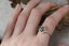 This custom fantasy engagement ring setting crafted by The North Way Studio features unfurling rustic leaves of hand-carved gold, rustling below a halo of natural white rose cut diamonds strewn about a uniquely chosen center stone. This variation of the Scrying Mirror ring is cast in 14 karat yellow gold and strewn about a teal rose cut sapphire. The engagement ring is shown worn on the index finger of a hand.