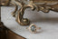 This elven fantasy engagement ring setting crafted by The North Way Studio features unfurling rustic leaves of hand-carved gold, rustling below a halo of natural white rose cut diamonds strewn about a uniquely chosen center stone. This variation of the Scrying Mirror ring is cast in 14 karat yellow gold and strewn about a teal rose cut sapphire. The engagement ring is shown resting on a marble table.