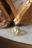 This custom antique fantasy engagement ring setting crafted by The North Way Studio features unfurling rustic leaves of hand-carved gold, rustling below a halo of natural white rose cut diamonds strewn about a uniquely chosen center stone. This variation of the Scrying Mirror ring is cast in yellow gold with a yellow rose cut diamond center and is shown resting on a marble table in front of a tipped over antique gold jewelry box.
