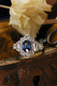 This fairytale custom heirloom engagement ring setting crafted by The North Way Studio features hand-carved gold leaves, rustling below a halo of natural white rose cut diamonds strewn about a uniquely chosen center stone. This variation of the ring is cast in 14 karat white gold with a deep blue oval sapphire center stone and shown resting on the edge of an antique wooden jewelry box.