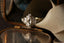 This antique inspired custom engagement ring setting crafted by The North Way Studio features a hand-carved leaves and a halo of natural white rose cut diamonds strewn about a uniquely chosen center stone. This variation of the ring is cast in 18 karat white gold with a natural elongated oval rose cut diamond center stone and shown resting in an antique glass and mirrored jewelry box.