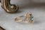 This custom fantasy engagement ring setting crafted by The North Way Studio features unfurling rustic leaves of hand-carved gold, rustling below a halo of natural white rose cut diamonds strewn about a uniquely chosen center stone. This variation of the Scrying Mirror ring is cast in 14 karat yellow gold and a teal rose cut sapphire. The engagement ring is shown facing the viewer's right and resting on a marble table.
