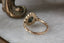 This heirloom engagement ring setting crafted by The North Way Studio features rustic leaves of hand-carved gold and a cluster of natural white rose cut diamonds set around and a uniquely chosen center stone. This variation of the Scrying Mirror ring is cast in 14 karat yellow gold and strewn about a teal rose cut sapphire. The engagement ring is shown facing away from the viewer.