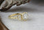 This custom fantasy heirloom engagement ring setting designed and crafted by The North Way Studio features two crescent diamonds and unfurling leaves of gold spun about a uniquely chosen center gemstone. This variation of the ethereal ring is shown cast in 18 karat green gold with a brilliant cut round diamond center. The ring is viewed close up as it rests facing the viewer's right on a marble table.