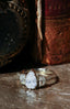 This custom heirloom engagement ring setting designed and crafted by The North Way Studio features two crescent diamonds and unfurling leaves of gold spun about a uniquely chosen center gemstone. This celestial fairy inspired ring is shown cast in 18 karat white gold with an opalescent fancy white pear diamond center. The ring is viewed facing slightly left while resting on an antique metal tray.