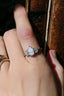 This customizable fantasy heirloom engagement ring setting designed and crafted by The North Way Studio features two crescent diamonds and unfurling leaves of gold spun about a uniquely chosen center gemstone. This ethereal fae engagement ring is shown cast in 18 karat white gold with an opalescent fancy white pear diamond center. The celestial ring is shown worn on the index finger of a hand resting on weathered books.