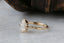 This custom heirloom engagement ring setting crafted by The North Way Studio features unfurling leaves and bark of carved gold, with petite claws to embrace a uniquely chosen center stone. This mythical ring is shown in 14 karat yellow gold with a pear cut diamond center. The fantasy engagement ring is shown close up resting on a marble table and facing the viewer's left, providing a view of the hand-carved details on the band.
