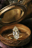 This customizable heirloom engagement ring setting crafted by The North Way Studio features unfurling leaves and bark of carved gold, with petite claws to embrace a uniquely chosen center stone. This magical ring is shown in 18 karat yellow gold with a large antique marquise old mine cut diamond center. The fairytale engagement ring is resting in an antique compact mirror tray.