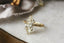 This customizable heirloom engagement ring setting crafted by The North Way Studio features unfurling leaves and bark of carved gold, with petite claws to embrace a uniquely chosen center stone. This mythical ring is shown in 18 karat yellow gold with a large antique marquise old mine cut diamond center. The fantasy engagement ring is shown close up resting on a marble table.