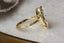 This custom heirloom engagement ring setting crafted by The North Way Studio features unfurling leaves and bark of carved gold, with petite claws to embrace a uniquely chosen center stone. This mythical ring is shown in 18 karat yellow gold with a large antique marquise old mine cut diamond center. The elven fantasy inspired engagement ring is shown close up resting on a marble table and facing the viewer's right, providing a view of the hand-carved details on the band.