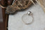 This custom fairytale heirloom engagement ring setting crafted by The North Way Studio features unfurling leaves and bark of carved gold, with petite claws to embrace a uniquely chosen center stone. This fantasy ring is shown in 14 karat white gold with a teal blue sapphire center. The alternative engagement ring is shown resting on a marble table viewed from above.
