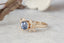 A yellow gold engagement ring set with a pale blue oval sapphire, framed by marquise and round diamond petals in a floral design along a carved bark-textured band, resting on pale marble beside a dried leaf.
