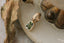 The Hag Stone Pendant in yellow gold: a deep green, rectangular, emerald-cut stone in a bezel edged with tiny beads, hanging from a rounded bail patterned with leaves. It rests on white marble beside a dried, curling leaf and an antique brass fitting.