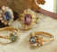 Four Secret Garden Collection engagement rings in yellow gold, set with a blue sapphire, a pink spinel, and mint and gray sapphires in floral diamond halos and carved leaf settings, arranged on cream linen.